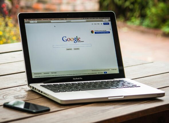 A MacBook Pro displaying Google Search on a wooden table outdoors, next to a smartphone.